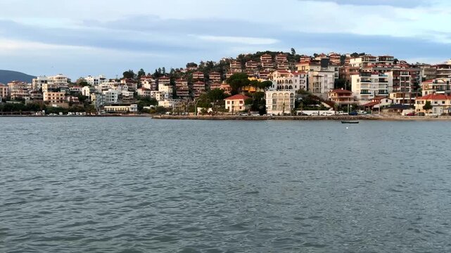 A scenic view of the coastal town of Gulluk, Turkey, with hotels and residential buildings terraced on a green hill overlooking the calm Aegean Sea under a soft, slightly overcast evening sky
