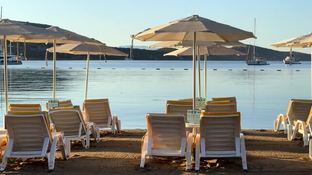 An empty sandy beach in Bitez, Bodrum, lined with neat rows of sun loungers and matching white umbrellas facing a calm, glassy blue bay beneath a bright morning sky