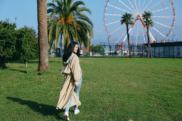 Fototapeta premium Woman strolling in a sunny park with a ferris wheel in the background, casual coat and sneakers, green grass and palm trees, daytime outdoor recreation scene