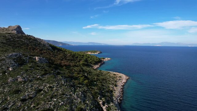 Majestic drone shot of the wild, rocky Moni island surrounded by the deep blue Aegean sea. Steep cliffs, green pine trees, and hills under a clear blue sky.