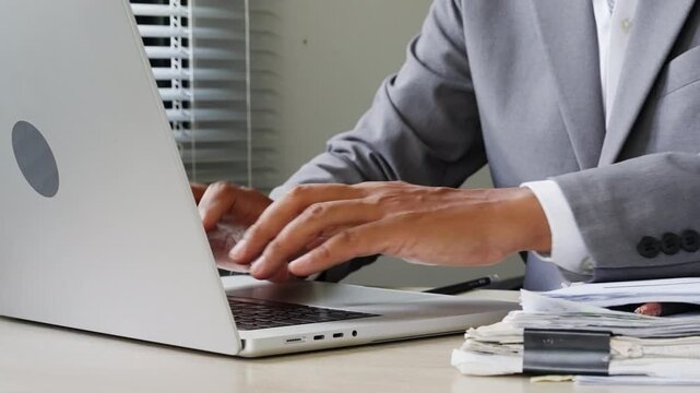 Business professional typing on laptop beside paperwork stack, representing digital workflow, office administration, document management, corporate reporting, productivity, and online business tasks.