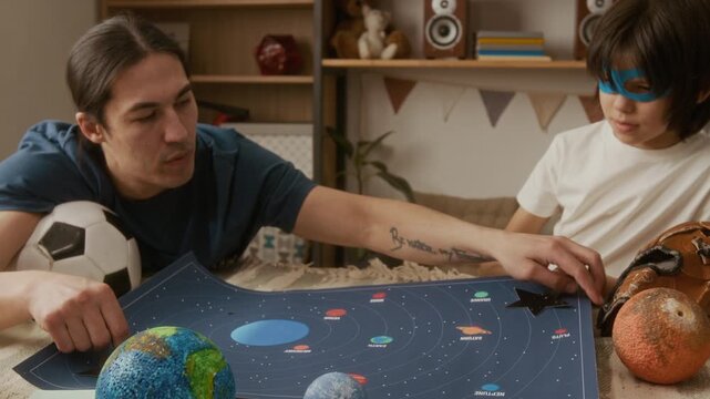 Father and young son studying solar system map on bed, placing astronaut toy on planets. Clip is perfect for homeschooling ads or educational product marketing