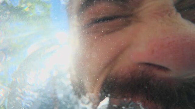 Bearded Man Dunks Face Underwater and Screams into Pool, Releasing Air Bubbles Against Bright Sun, Clouds and Tree Leaves in Distorted Upward View Dramatic Summer Aquatic Close Up Scene Shot
