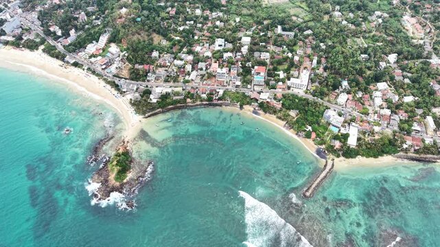 Aerial view of Parrot Rock and the turquoise waters of Mirissa Beach surrounded by lush tropical greenery and coastal buildings in Mirissa, Southern Province, Sri Lanka.