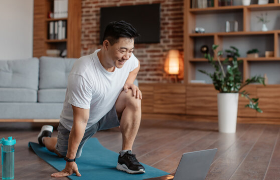 Sporty asian mature man exercising at home, stratching his legs, warming up before training and using laptop, watching fitness video on Internet or having online fitness class at home