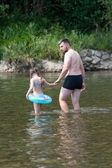 Fototapeta premium A father and daughter wade in a shallow river, the girl holding a small inflatable ring.