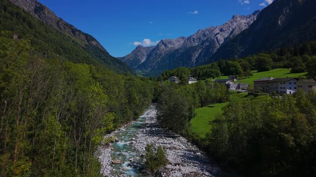 Drone view flying over pristine river and lush green forest with swiss alps in background in Bregaglia Valley, Switzerland. Aerial forward