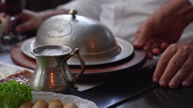 Close-up of a waiter serving a traditional meal under a hammered metal cloche at a restaurant. Includes a metal mug with foamy ayran drink and side appetizers in a cinematic setting.