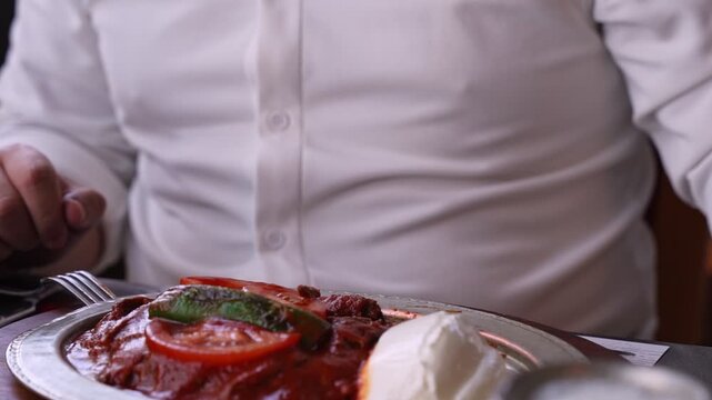 Close-up of hands tearing fresh pita bread over a traditional Turkish Iskender kebab platter with meat, tomato sauce, and yogurt in a cinematic restaurant setting.