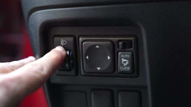 Close-up of a finger turning the headlight adjustment dial on a vehicle dashboard. Cinematic shot showing automotive interior controls, side mirror buttons, and auto close feature.