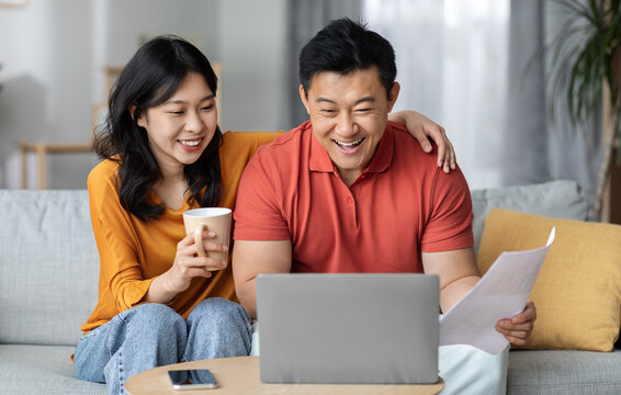 Smiling asian spouses paying bills online while staying home, sitting on couch, using laptop, happy chinese man holding papers and banking on Internet, cheerful woman drinking tea, copy space
