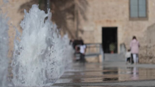 Super slow motion of the water jets in the plaza in front of the Altamira Palace. Out-of-focus family heading towards the palace entrance. Elche, Spain.