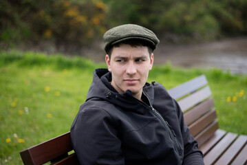 Fototapeta premium A guy looks with envy and a smile at young couple sitting on a bench not far from him and guy misses his girlfriend. A pensive young man wearing a cap and jacket sits on a wooden park bench