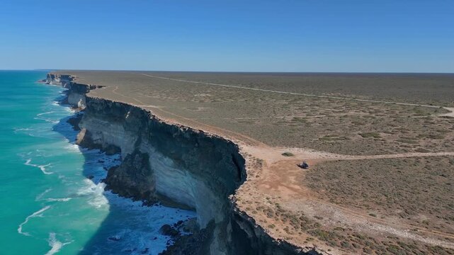 Drone shot orbiting a parked car from the ocean side near the Bunda Cliffs, revealing endless sheer cliffs with deep blue sky and ocean