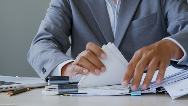 Businessman sorts papers at desk. Hands grip stacks of documents with focus. Office environment suggests planning or review. Scene conveys diligence and organization