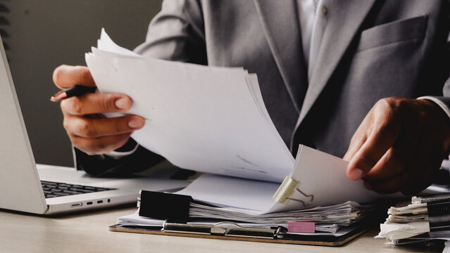 Businessman reviews documents near laptop. Focused, serious demeanor suggests decision-making. Desk holds papers, pens, and office supplies. Professional setting implies productivity and strategy