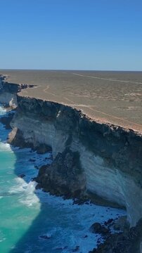 Vertical drone shot panning from the Bunda Cliffs toward the ocean, revealing white waves and a distant horizon