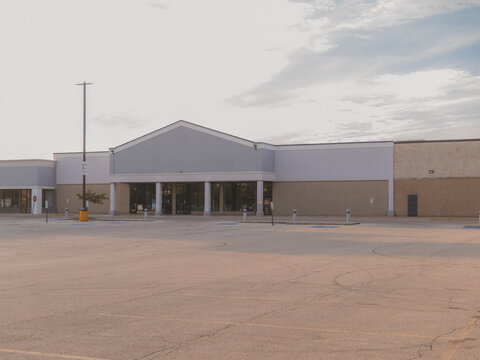 An empty abandoned big box storefront building with a blank gabled sign space and a vacant asphalt parking lot under a hazy sky