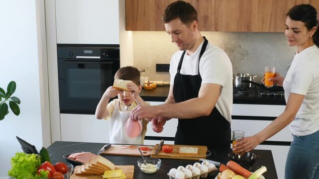 Father and young boy make sandwiches in a contemporary kitchen, surrounded by fresh vegetables and ingredients on the countertop