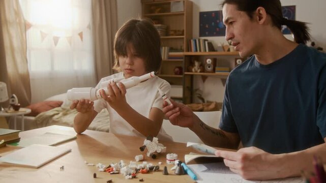 Asian young adult man and multiethnic child building toy space rocket from plastic blocks at table. Clip is perfect for promoting creative toys, or family bonding campaigns