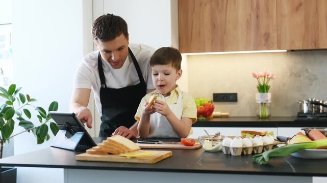 Father and son engage with a tablet in a modern kitchen, preparing food with various ingredients and utensils visible on the countertop