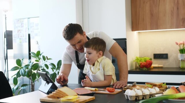 Father and son engage with a tablet in a contemporary kitchen, surrounded by food ingredients including bread, ham, and tomato on the countertop