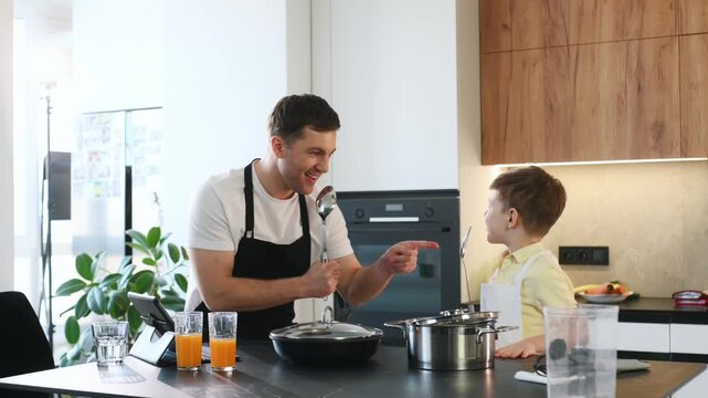 Caucasian man and boy interact playfully in a kitchen, using utensils and cookware, with fresh vegetables and modern appliances in the background