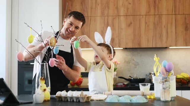 Father and son wearing bunny ears prepare for Easter celebration in a modern kitchen with colorful decorations and festive elements on the countertop