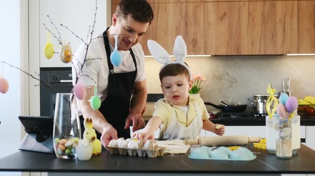 Father and young son hang colorful eggs on a decorative tree in a contemporary kitchen, surrounded by Easter-themed decorations and spring flowers
