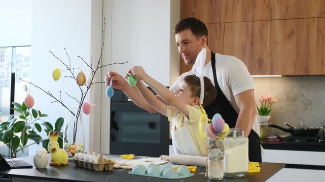 Father and young boy in bunny ears engage in Easter preparations in a bright kitchen, surrounded by colorful decorations and festive items on the counter