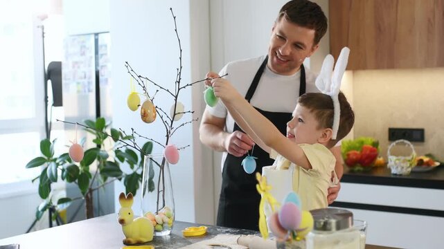 Father and young son hang colorful eggs on a decorative tree in a contemporary kitchen, surrounded by Easter-themed decorations and spring flowers