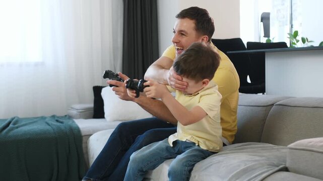 Father and young boy engaged in video gaming on a couch, with a contemporary kitchen area and dining chairs in the background