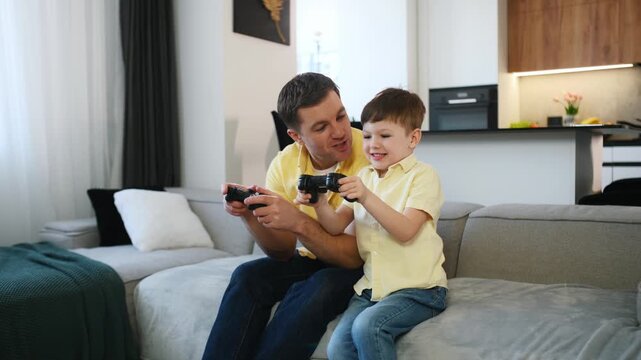 Caucasian man and boy joyfully celebrate while sitting on a sofa, both in yellow shirts, with a video game controller in a contemporary living room