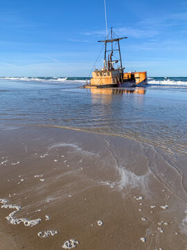 A weathered shipwreck rests in the shallow surf of North Carolina&rsquo;s Outer Banks, surrounded by gentle waves, reflective wet sand, and expansive coastal skies. 