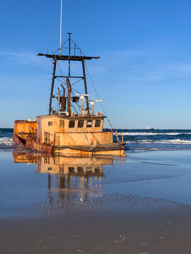 Abandoned shipwreck reflected in wet sand along the Outer Banks coastline, with Atlantic waves and blue skies creating a striking maritime scene.
