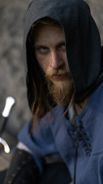 A rugged man with a red beard and piercing gaze stares intently while wearing a hooded tunic.