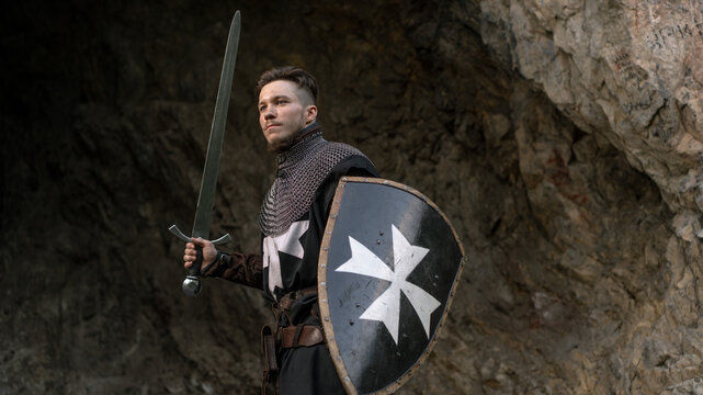 A knight stands confidently with a sword and shield against a rocky backdrop.