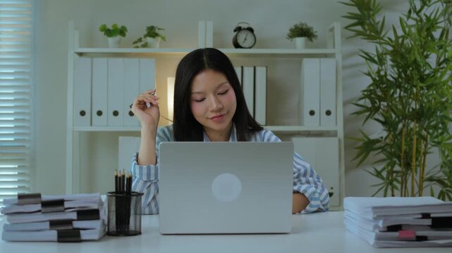 Asian Woman Sitting at Desk Holding Pen and Thinking While Working on Laptop. Focused Professional Planning and Decision Making Moment.