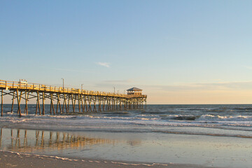 Fototapeta premium The Atlantic Beach Fishing Pier extends into the Atlantic Ocean as evening light reflects across wet sand and rolling waves along the North Carolina coast.