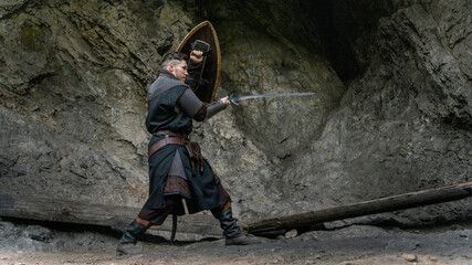 A medieval warrior stands ready with sword and shield against a rocky backdrop. © horimono