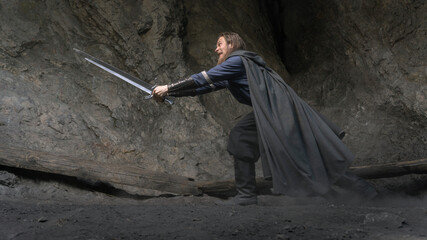 A rugged warrior stands poised with a gleaming sword amidst a rocky cavern, dust swirling around his feet. © horimono