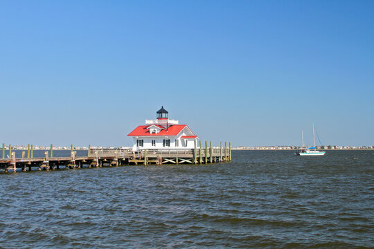 The Roanoke Marshes Lighthouse stands above the waters of Albemarle Sound in Manteo, North Carolina, while a sailboat drifts across the calm coastal landscape.