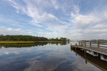 Fototapeta premium A peaceful coastal marsh in Croatan National Forest reflects sweeping clouds and surrounding pine forests, with a wooden observation deck extending over calm waters.