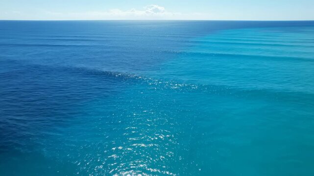 Expansive vista of calm, shimmering azure ocean waters stretching towards a distant, hazy horizon under a bright, cloudless sky, showcasing the beautiful contrast of two distinct, luminous blue