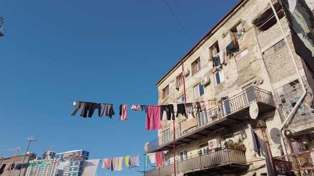 Low-angle view of laundry drying on clotheslines strung between old, weathered apartment buildings in a poor neighborhood, showing a stark contrast with modern skyscrapers in the distance