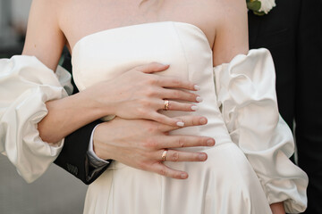 Obraz premium Romantic wedding detail. Closeup of a newlywed couple during an intimate embrace. The groom stands behind the bride and gently holds her waist with hands displaying their gold wedding rings.