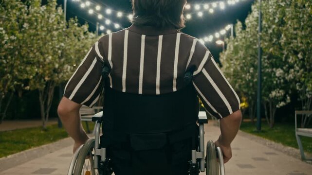 Man using wheelchair rolls forward along paved walkway while looking aside under warm lights. Evening garden, benches, and flowering trees suggest independence, freedom, and peaceful outdoor leisure.