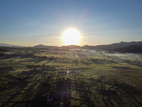 Drone view of sunrise over Pronojiwo farmland with morning mist, rolling hills, and agricultural fields illuminated by golden sunlight in East Java, Indonesia.