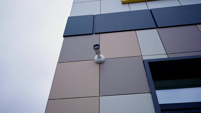 Geometric wall tiles contrast with sleek black window. Surveillance equipment monitoring perimeter of modern facility under overcast sky