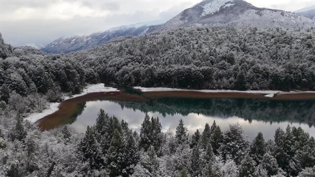 Snow covered Andean forest surrounding Lago Guillelmo in R&iacute;o Negro, Argentina with reflective glacial waters and mountainous Patagonian landscape, slow forward drone flyover shot